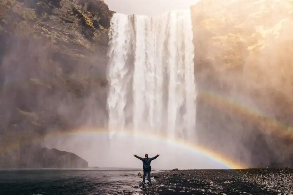 viajante levanta as mãos na frente de uma cachoeira com um arco-íris