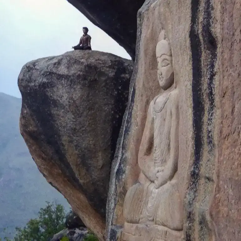Um homem sentado na posição de ioga de lótus sentado em uma rocha no final de um penhasco com um Buda esculpido no penhasco em primeiro plano