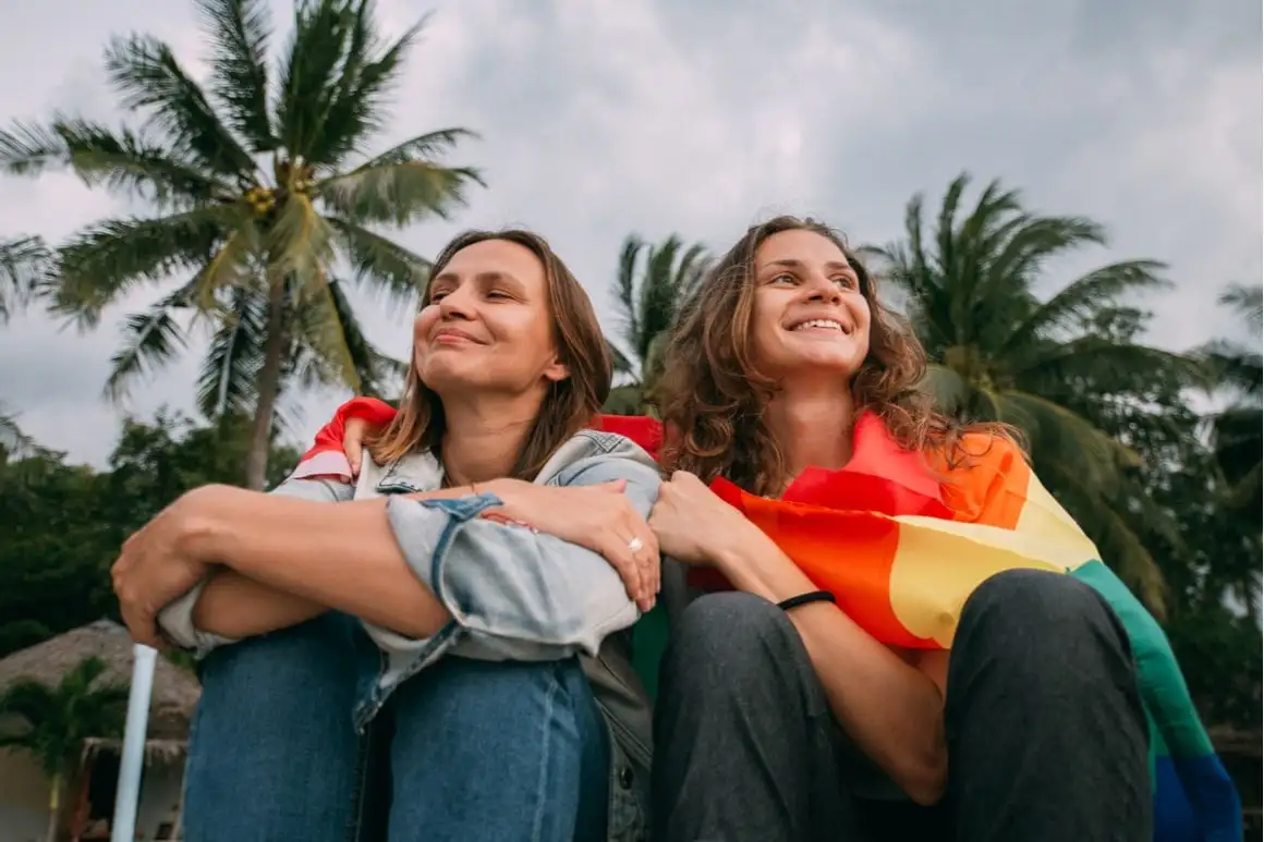 Duas garotas sentam-se lado a lado com a bandeira do orgulho LGBTQ enrolada nelas.