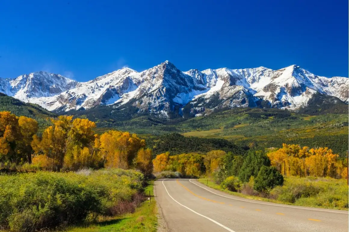 Montanhas cobertas de neve e árvores de outono estão ao longe. Uma estrada reta sem carros está em primeiro plano.