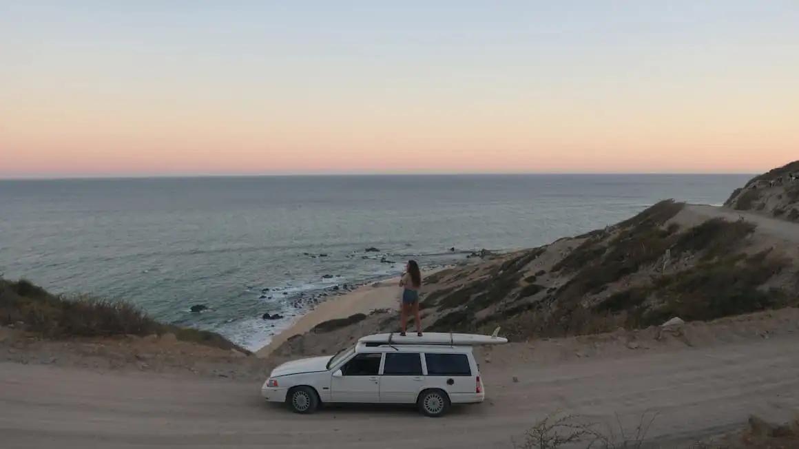 une fille debout au sommet d'une voiture blanche transformée en camping-car alors qu'elle regarde le coucher du soleil et l'océan