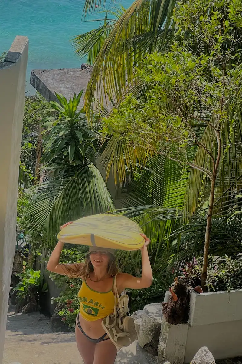 une fille tenant une planche de surf sur sa tête alors qu'elle monte les escaliers avec la plage et les palmiers en arrière-plan