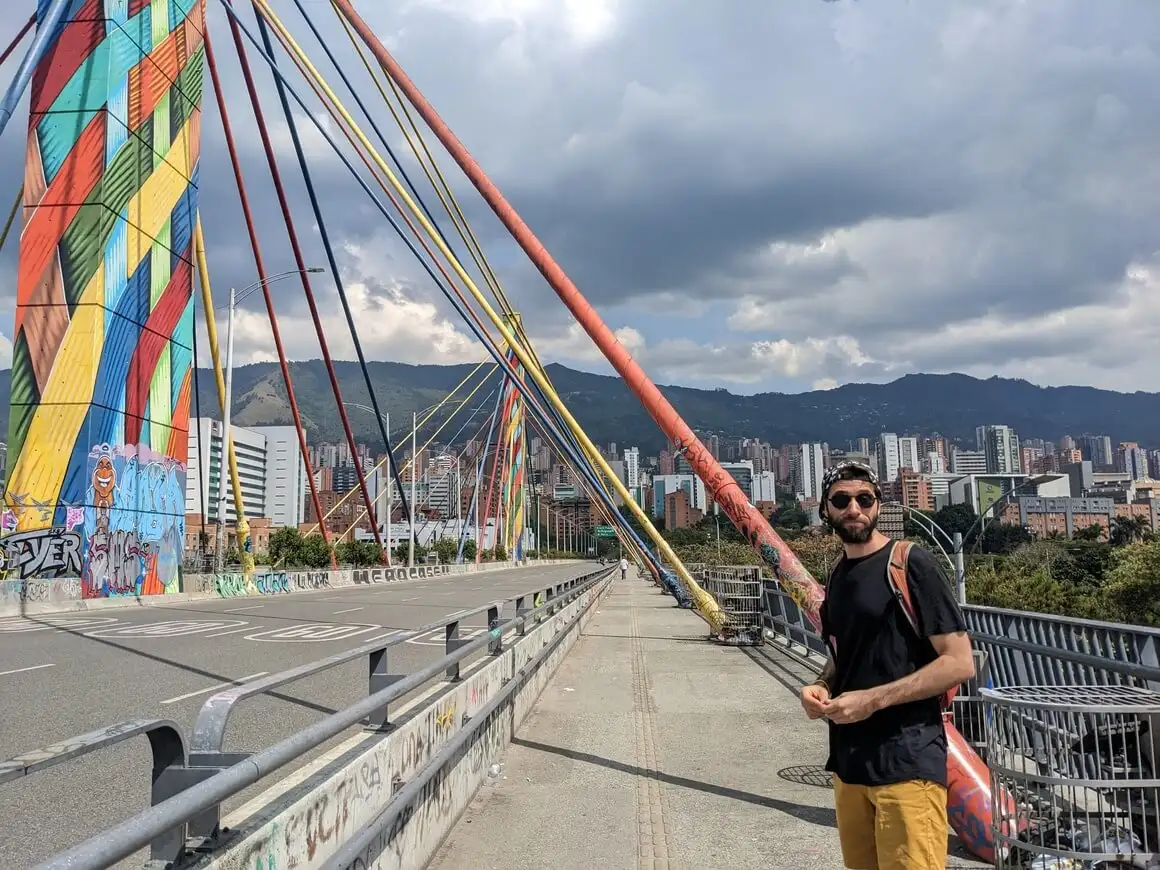 Homme avec un t-shirt noir, une casquette et des lunettes de soleil, sur le pont coloré de la rivière Medellin