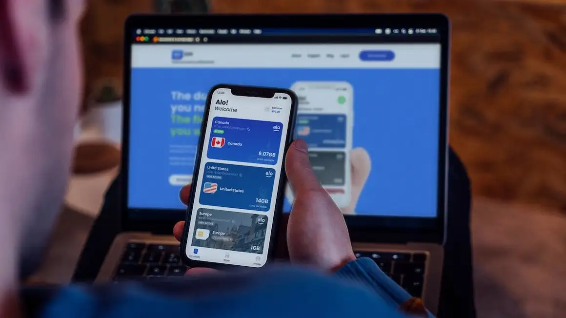 personne tenant un téléphone avec l'application aloSIM ouverte avec le site Web ouvert sur son ordinateur portable