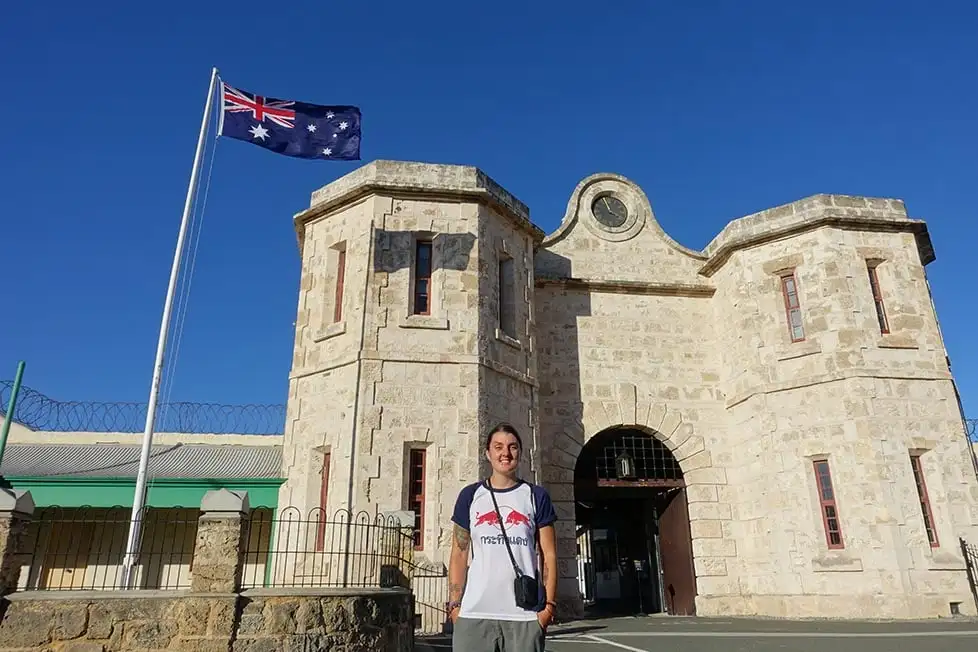 Une personne debout devant la prison de Fremantle, en Australie occidentale. Derrière eux se trouvent les grandes portes d'entrée en pierre et un drapeau australien.