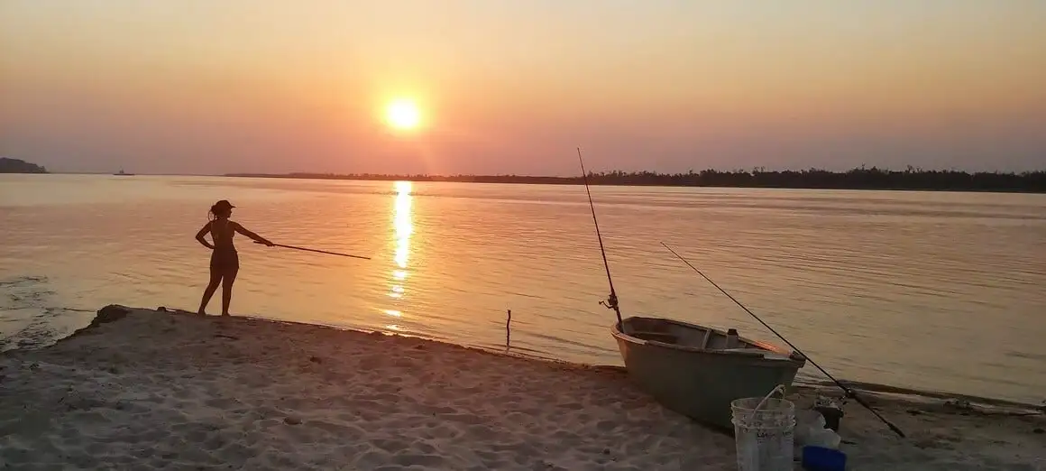 Silhouette d'une femme pêchant dans une rivière devant un coucher de soleil rouge-orange