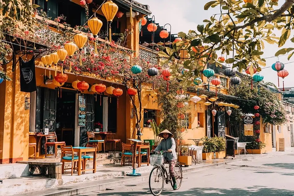 Une personne sur un vélo avec un chapeau vietnamien passant devant une maison jaune avec des lanternes colorées à Hoi An, Vietnam