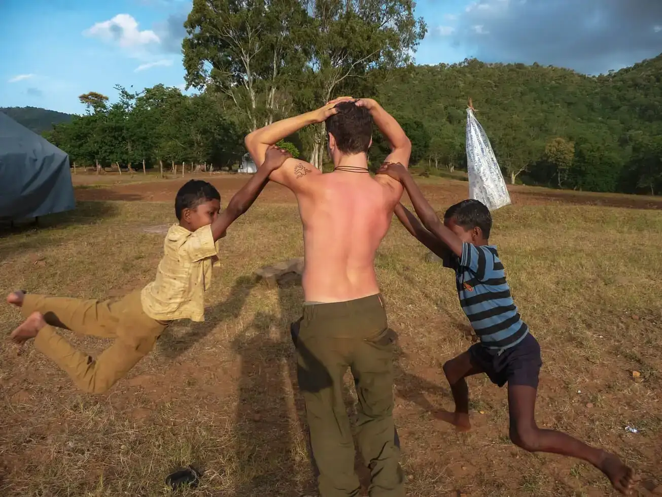 homem sem camisa fazendo voluntariado na Índia rural com duas crianças balançando nos braços