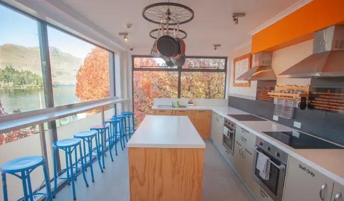 Cuisine spacieuse et ensoleillée avec cuisinières, îlot et rangée de tabourets près d'un comptoir côté fenêtre donnant sur l'eau et les montagnes à l'Absoloot Hostel à Queenstown, Nouvelle-Zélande