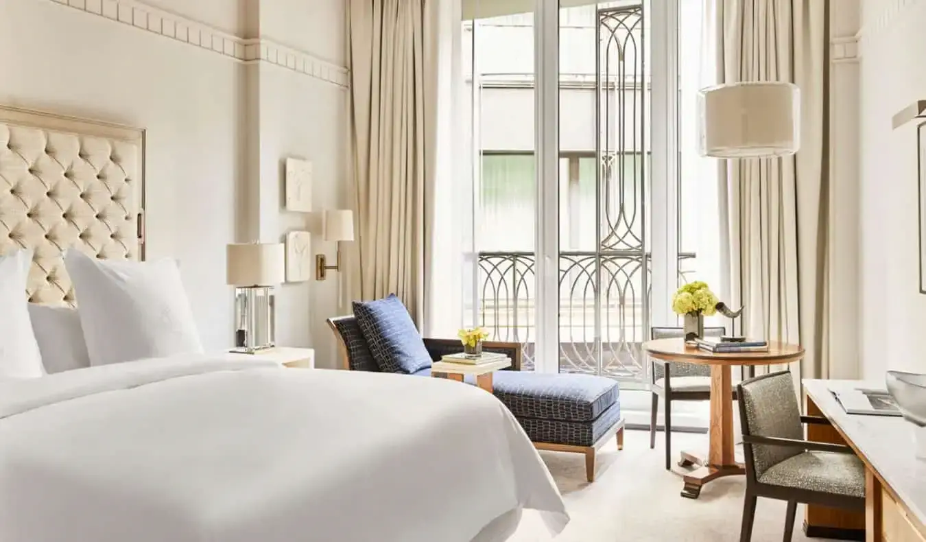 Une luxueuse chambre d'hôtel de couleur crème avec un grand lit, une chaise longue bleue près de la fenêtre ouverte et un bureau au Four Seasons de Madrid, en Espagne