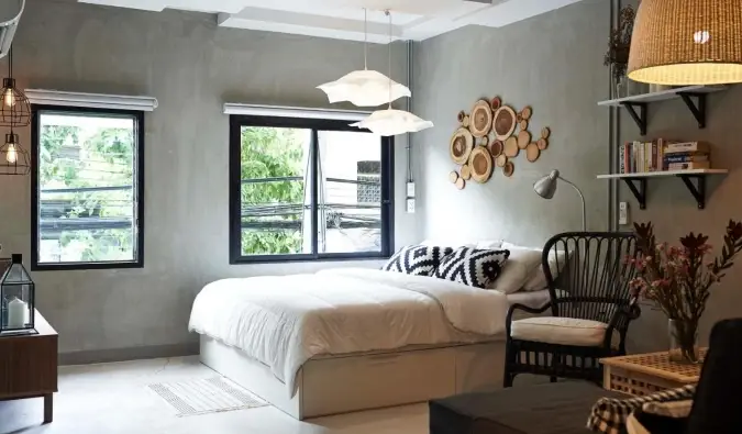 Un lit double dans une chambre d'hôtel avec de grandes fenêtres, des fenêtres en béton brossé et des accents en bois sur les murs de l'hôtel House of Phraya Jasaen à Bangkok, en Thaïlande