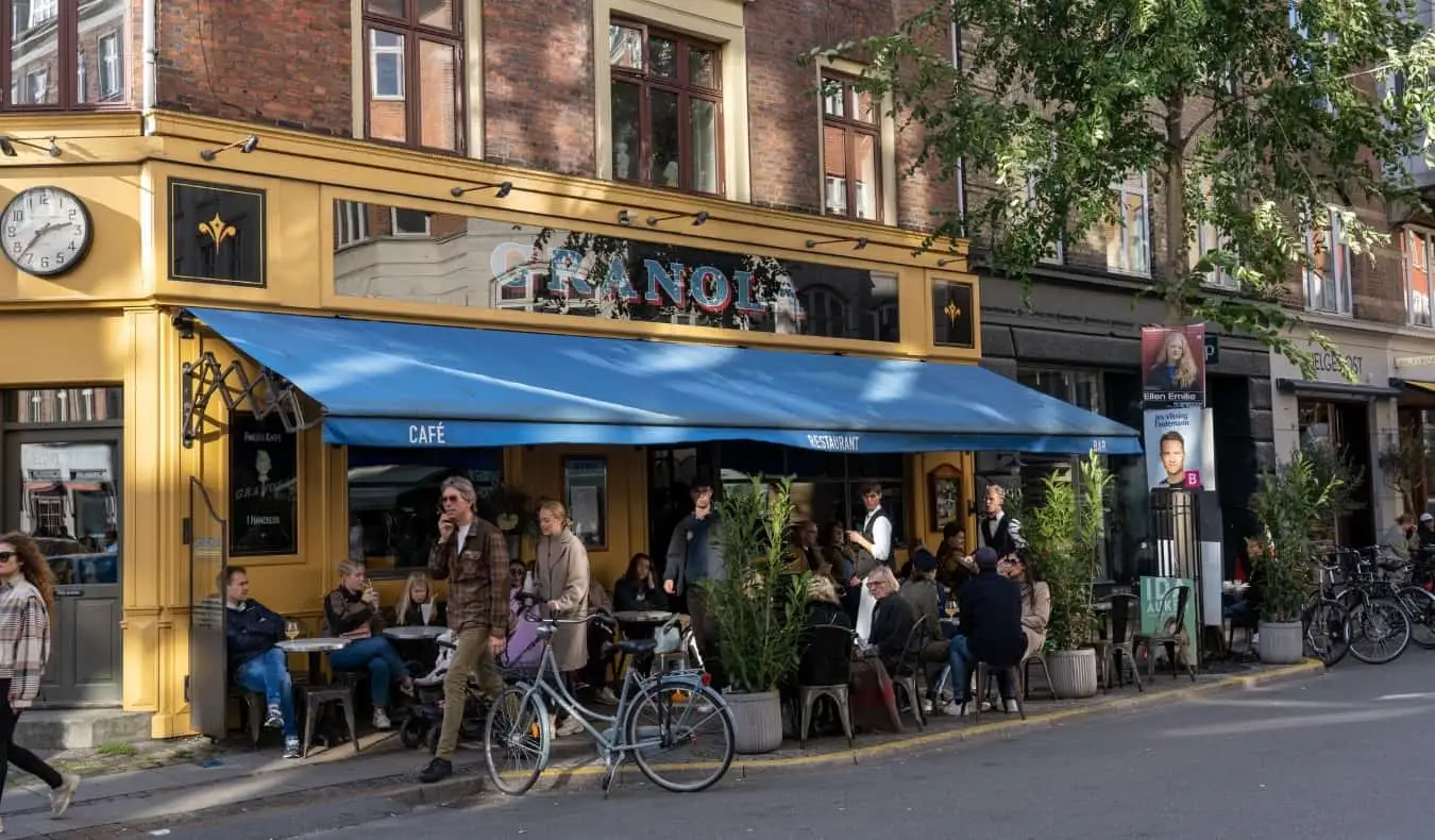 Passants devant des personnes assises dans une terrasse de café dans une rue verdoyante du quartier de Vesterbro à Copenhague, Danemark