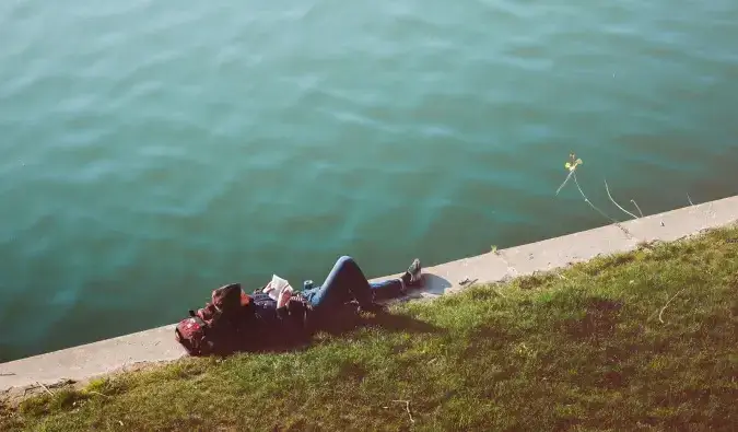 Ein preisbewusster Reisender liegt im Gras und liest ein Buch am Wasser