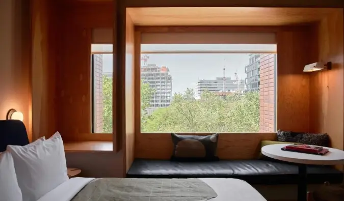 Uma cama em frente a uma janela panorâmica em uma parede com painéis de madeira, com um banco na frente dela no Ace Hotel em Toronto, Canadá