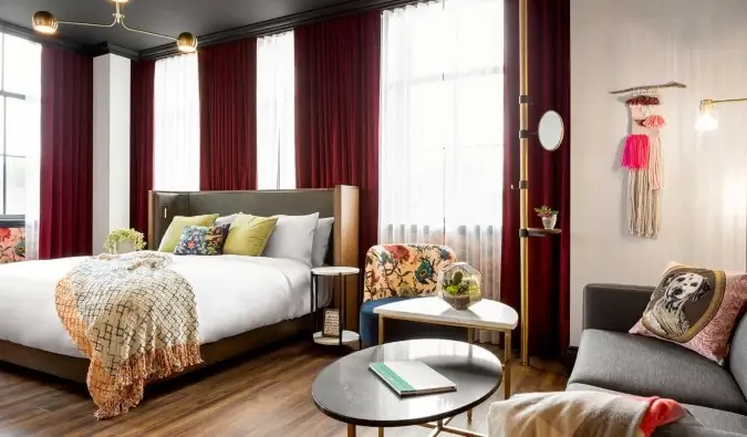 Um grande quarto com uma cama em frente a janelas do chão ao teto forradas com ricas cortinas vermelhas e um sofá e uma mesa ao lado no The Broadview Hotel em Toronto, Canadá