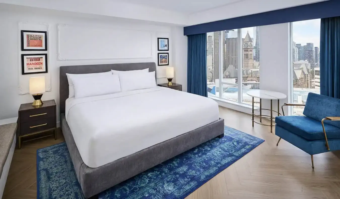 Uma cama king-size com porta-retratos pendurados nas paredes de ambos os lados e grandes janelas do chão ao teto com vista para a paisagem urbana de Toronto, Canadá, no Pantages Hotel Downtown