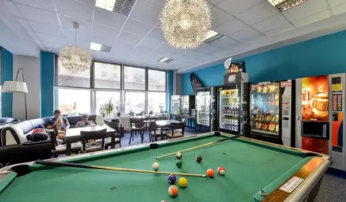 Salle commune avec table de billard, distributeurs automatiques, tables et personnes se prélassant sur des canapés au PLUS Hostel à Prague.