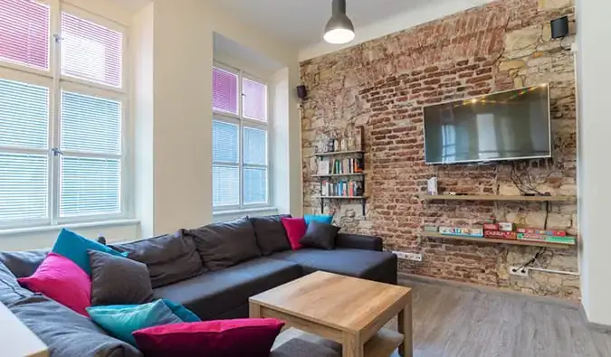Salle commune avec canapé en L, murs en briques apparentes, télévision et étagères avec livres et jeux à l'auberge The RoadHouse à Prague.