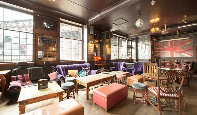 Salle commune confortable et spacieuse avec de nombreuses tables et chaises en bois, des canapés violets et un grand drapeau britannique sur le mur du Walrus Hostel, Londres