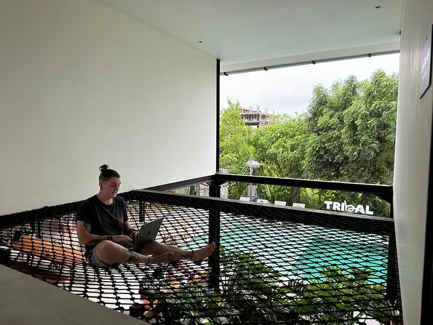Nic trabalhando em seu laptop em uma rede de carga em um albergue com piscina ao fundo.