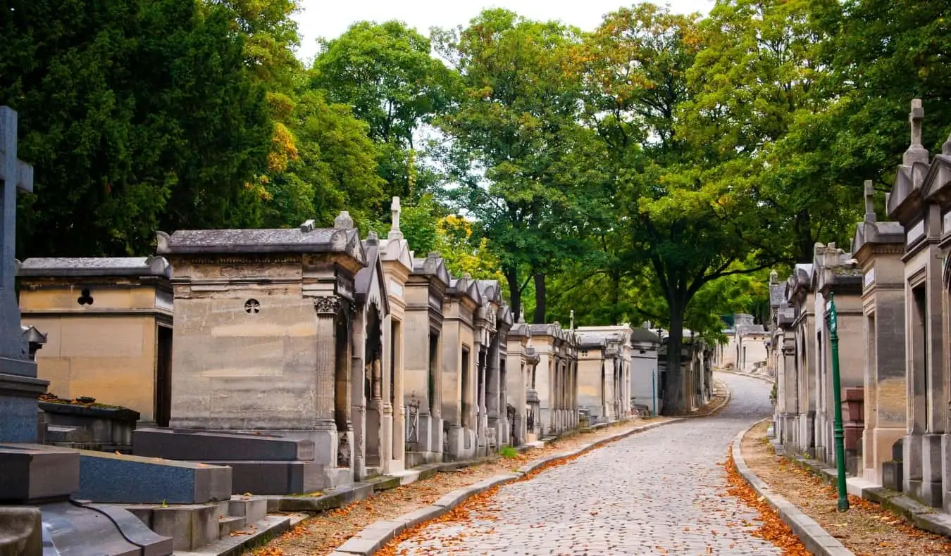 Mausoléus alinhados ao longo de um caminho de paralelepípedos repleto de folhas de laranja no Cemitério Pere Lachaise, em Paris, França
