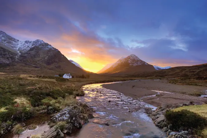 Foto do pôr do sol atrás das montanhas em Glencoe, Escócia, com uma casa no meio do terreno