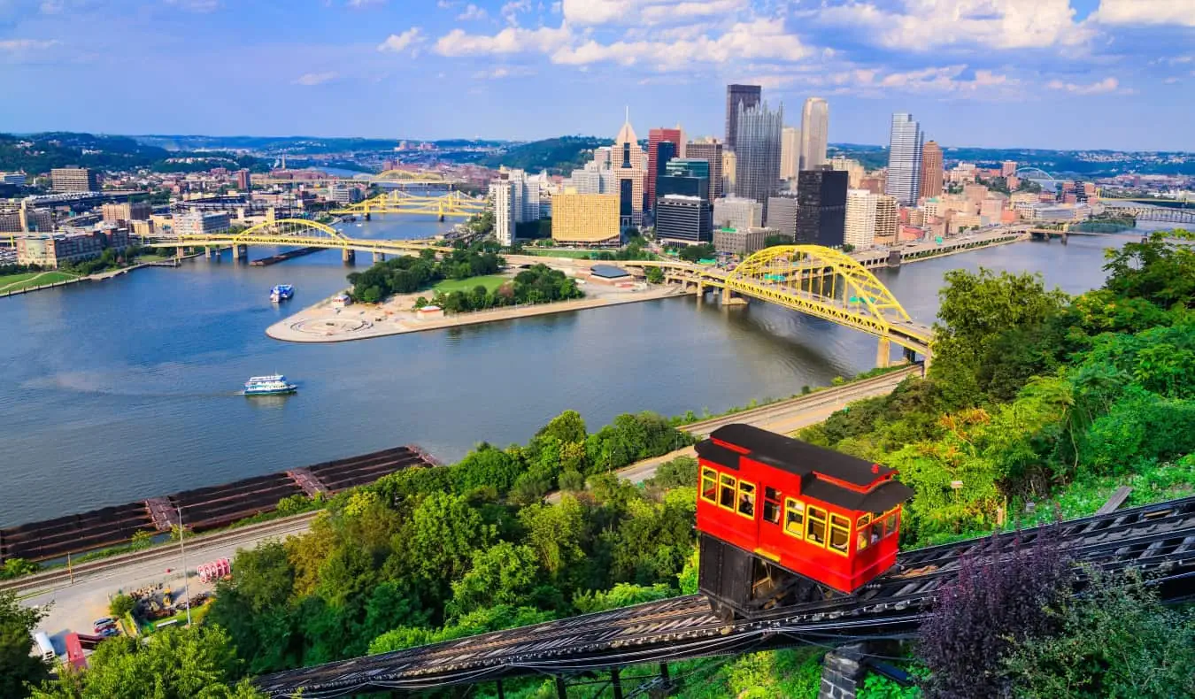 Um funicular vermelho subindo a montanha em primeiro plano com o horizonte de Pittsburgh, PA, com suas muitas pontes que atravessam o rio ao fundo